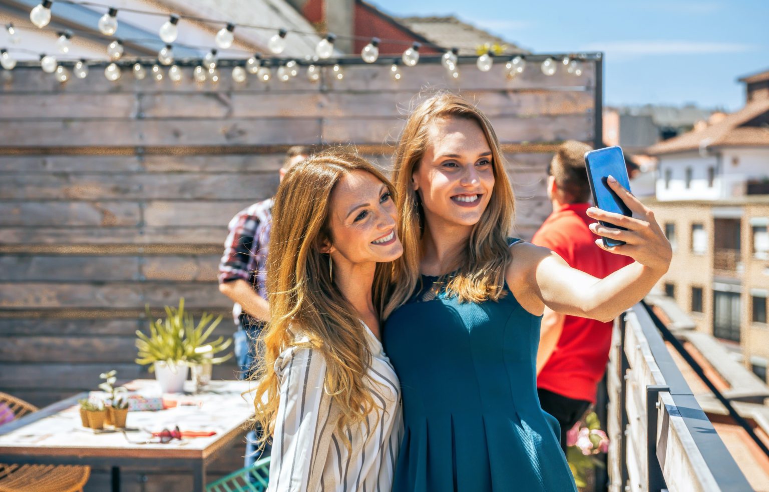 Portrait of two smiling female friends taking a selfie with phone in office rooftop on a summer day while their colleagues talking on background